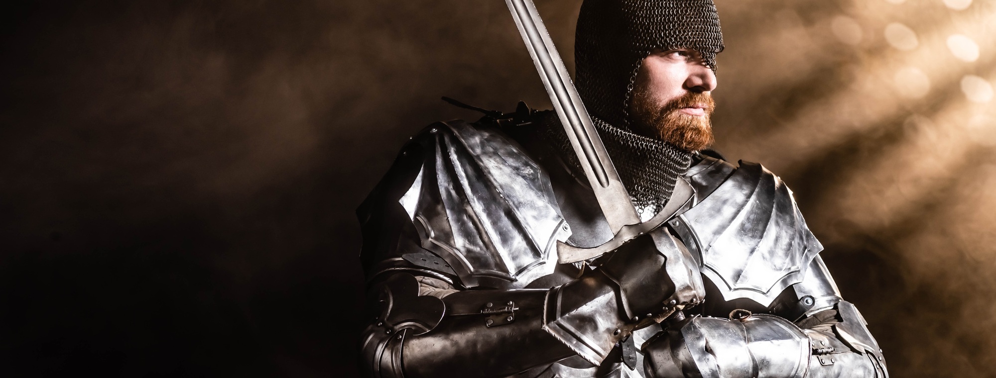 panoramic shot of handsome knight in armor holding sword on black background